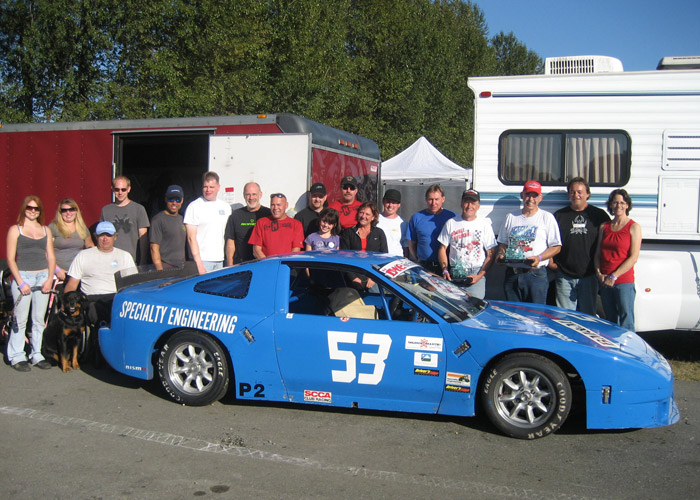18 people and a dog pose behind side view of /53 blue Nissan 240SX racing car